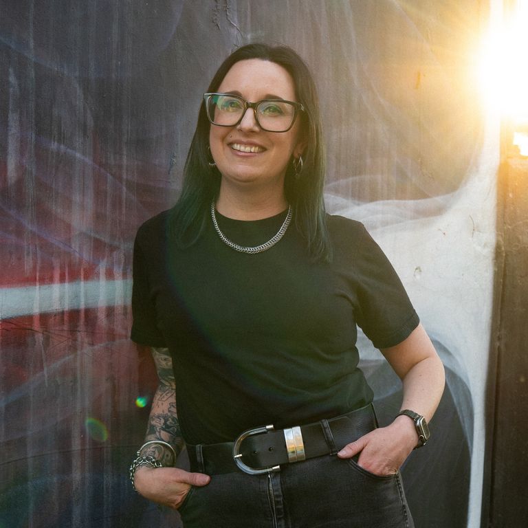 Portrait of Anita Decoster wearing glasses and a black shirt against a painted wall outdoors.